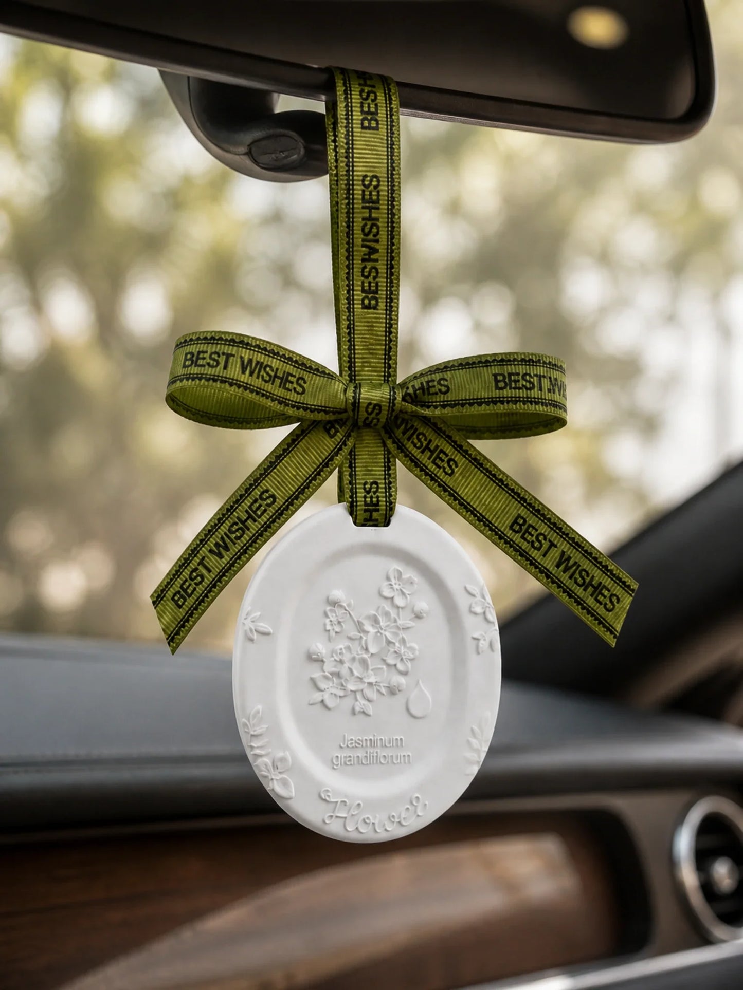 White Jasmine medallion hanging beneath a rearview mirror with its original olive BEST WISHES ribbon
